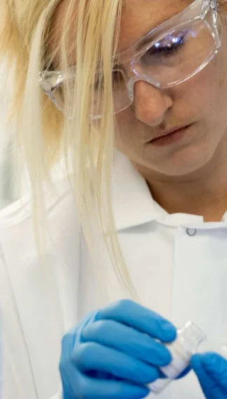 A female scientist in a lab coat and safety goggles examining a test tube closely.