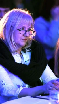 A woman with blonde hair and glasses, wearing a black scarf and white shirt, sits attentively in a blue-lit environment.