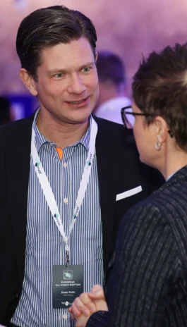 Two people in conversation at an event. One person wears a striped shirt and conference badge, the other has short hair and glasses.