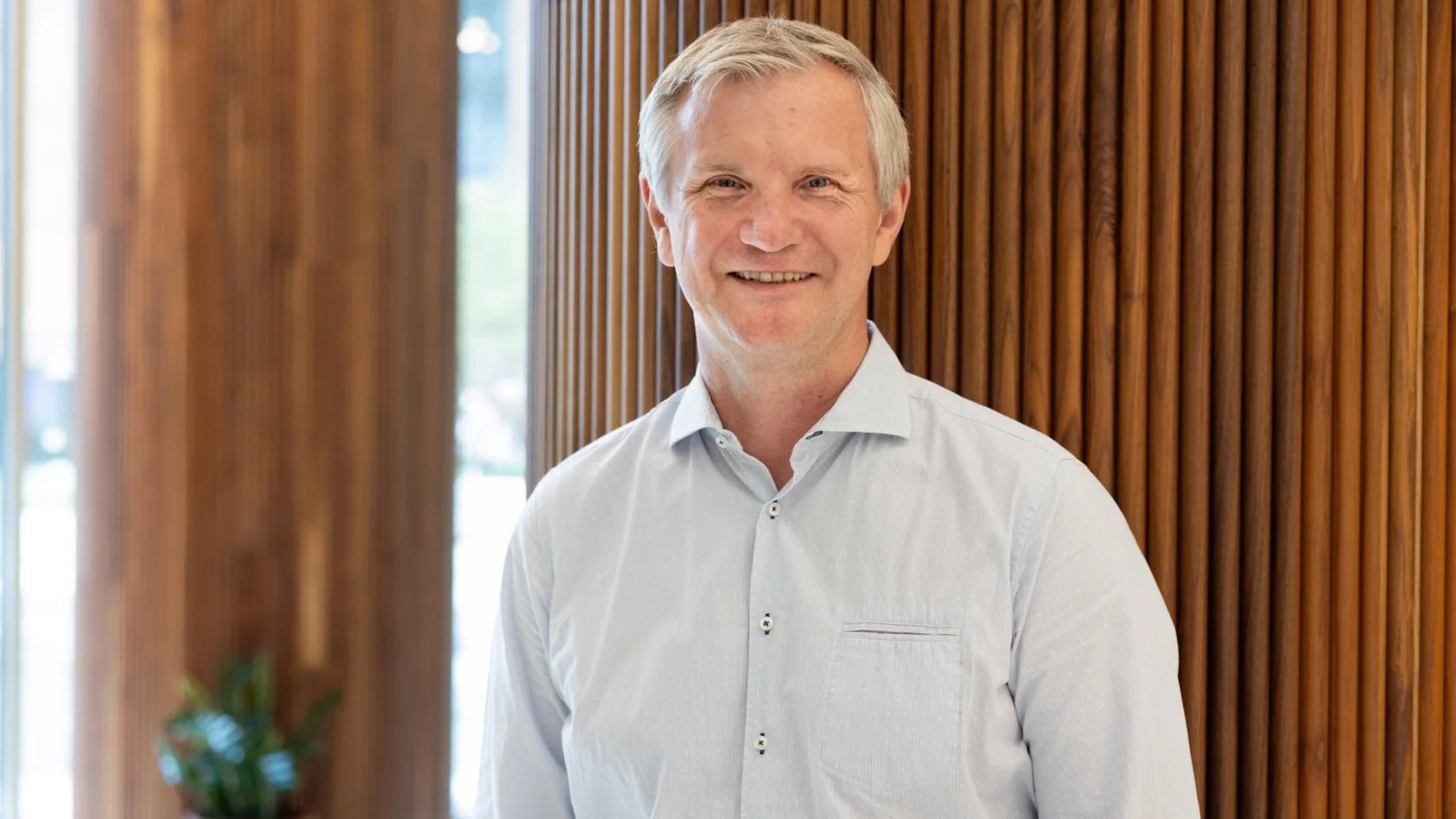 A smiling man in a light blue shirt stands against a wooden wall backdrop.