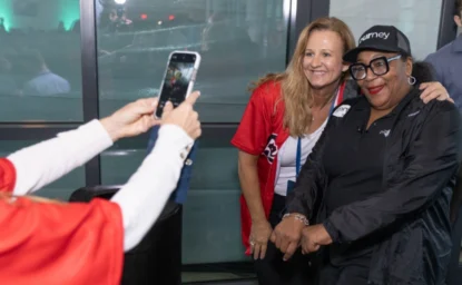 Two women pose for a photo taken by another person indoors. One woman wears a red jersey, and the other is dressed in black, including a cap and glasses.